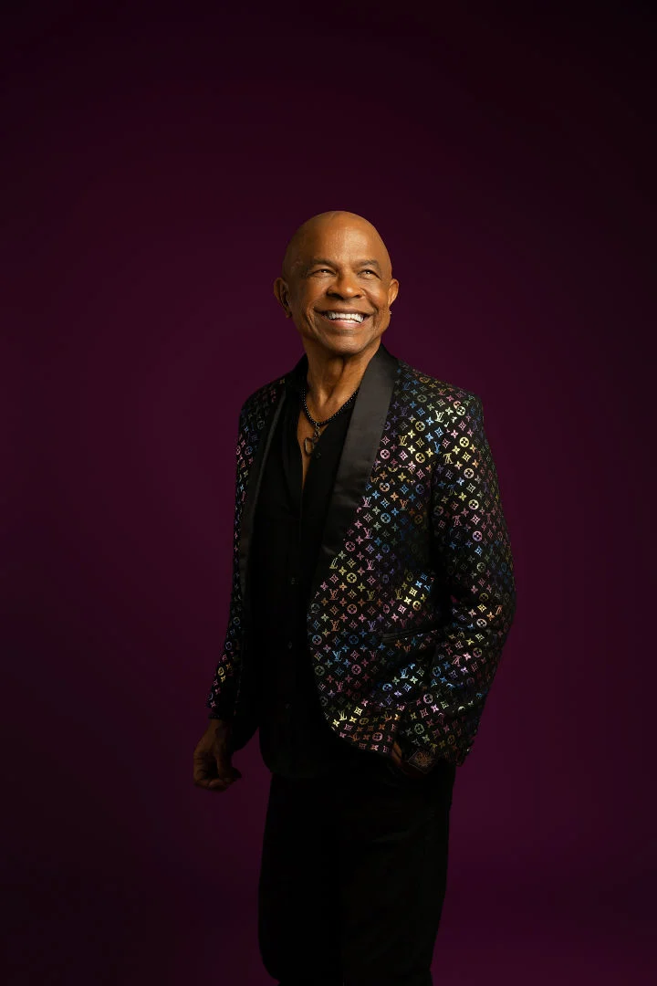 Francky Vincent souriant, en veste à motifs multicolores sur fond violet.
