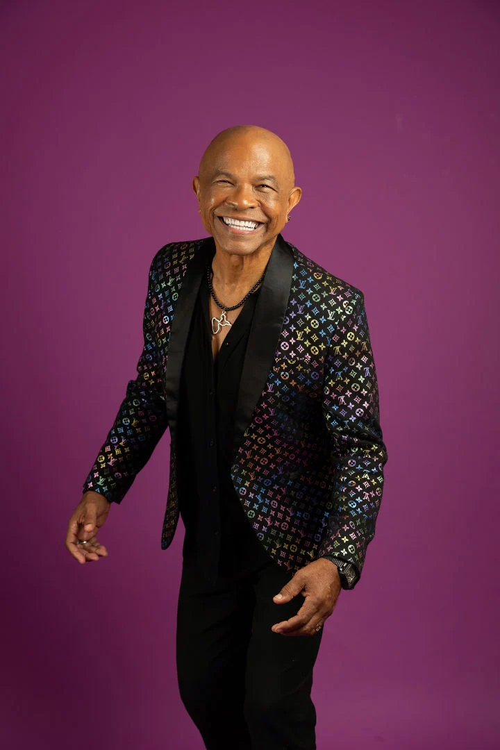 Portrait de Francky Vincent souriant face à l’objectif, en chemise noire et veste à motifs sur fond violet.